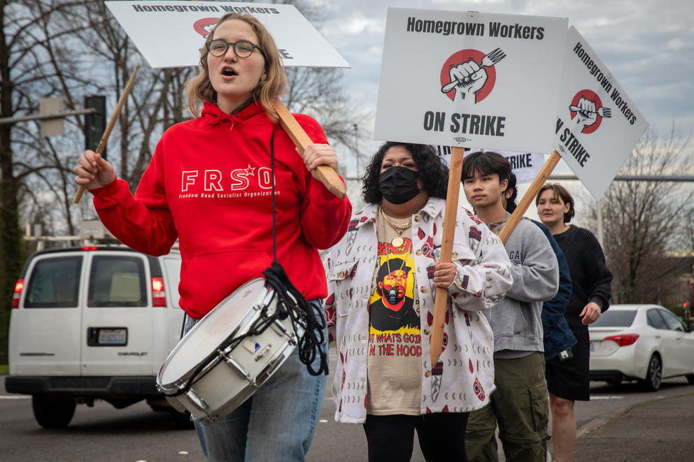 A person marches with a snare drum and picket sign, leading a handful of workers on strike.