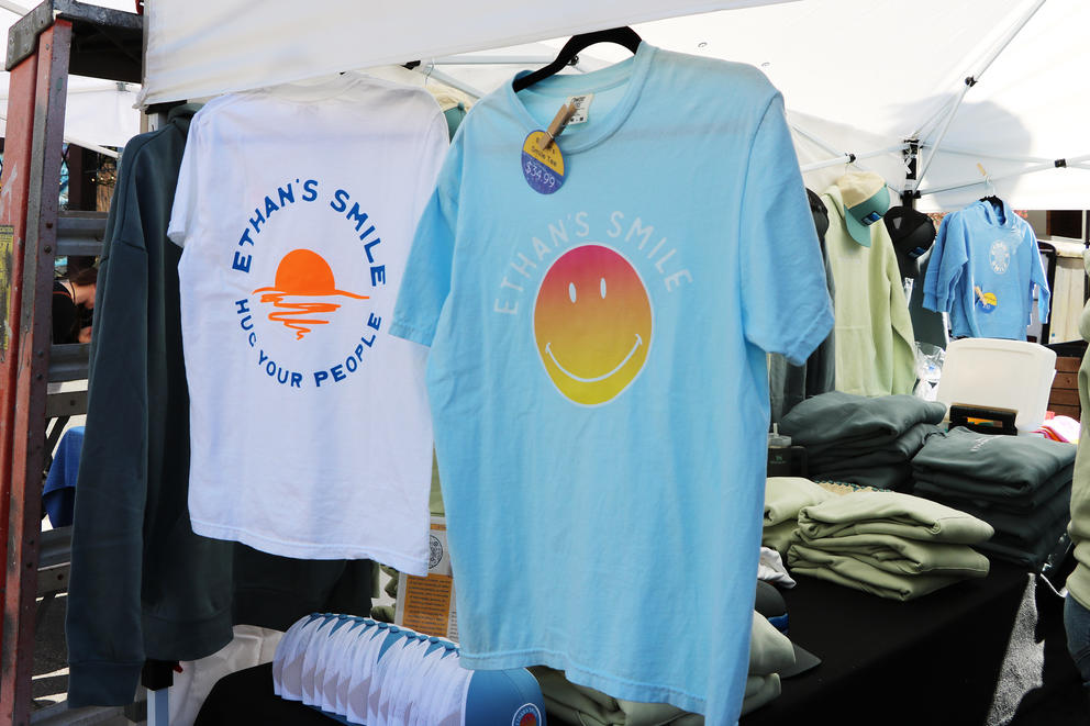 A white shirt hangs next to a blue shirt in a market tent. They both say "Ethan's Smile."