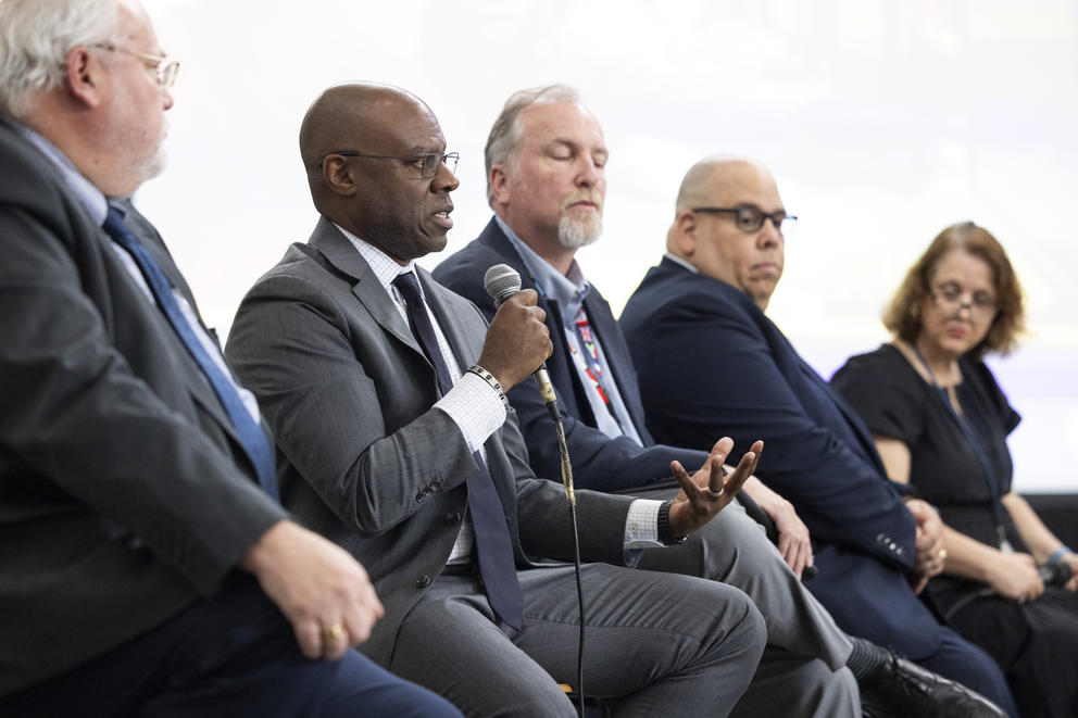 Four men and a woman sit on stools, one man is speaking into a microphone