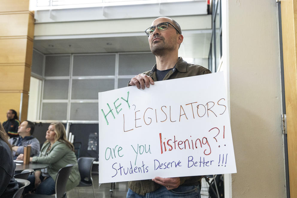 A man stands with a sign that reads "Hey, legislators are you listening?! Students deserve better!!!