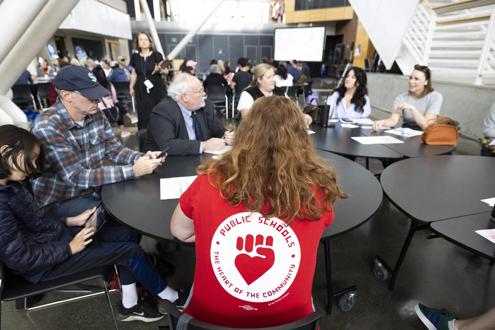 A woman is seen from the back at a table with other people. She is wearing a red shirt that reads "public schools, the heard of the community"