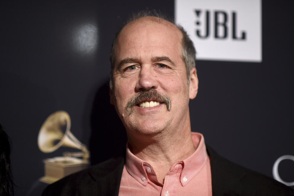 A person with a moustache smiles in front of a backdrop that has the logo of the Grammy awards.