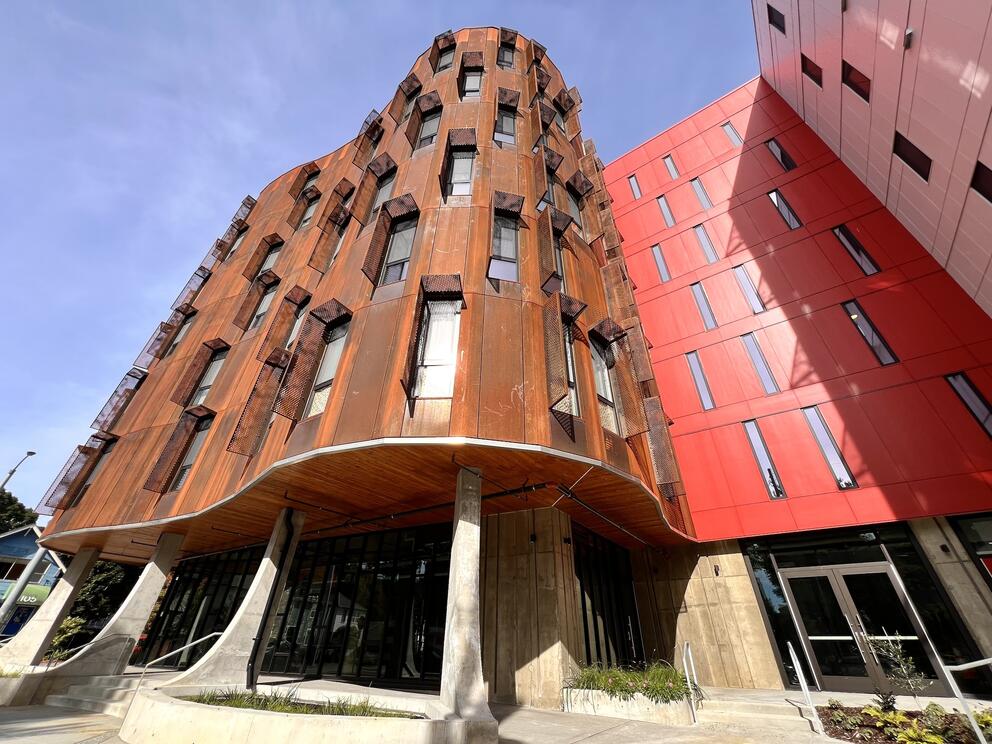 exterior of a windowed building with a curving brown facade set on arced supports, next to a red rectilinear building