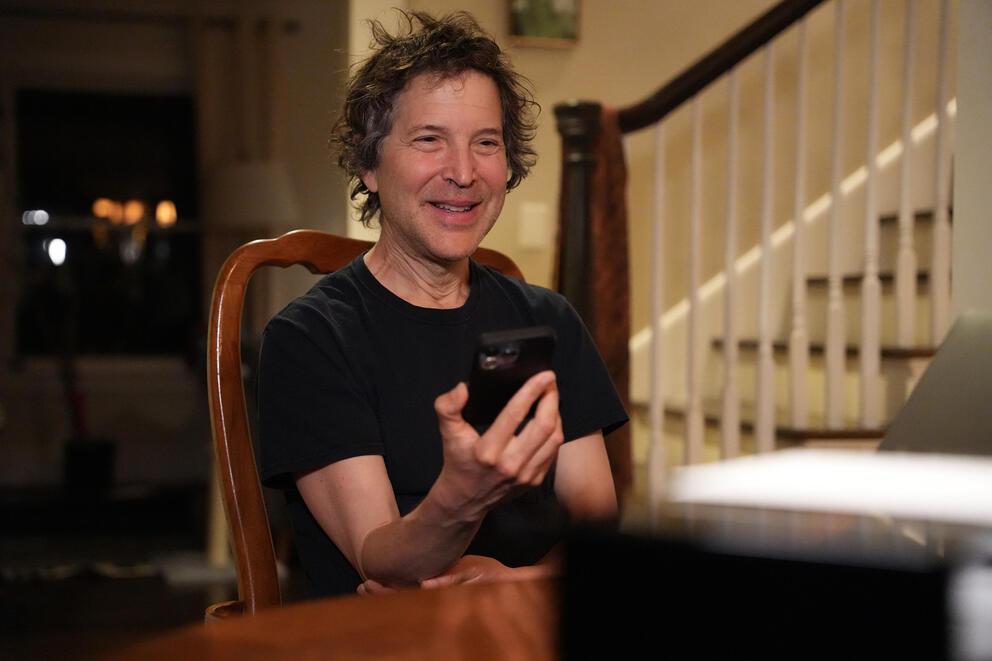 a 62 year old man in a black t shirt sitting at a kitchen table in his home smiles while holding his smart phone in front of him