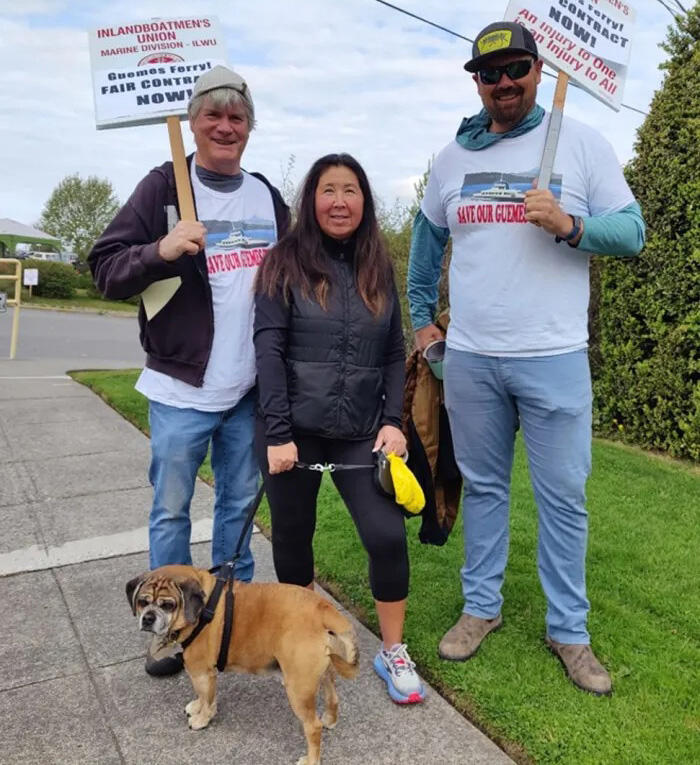 Three people pose for a photo outside holding signs that say, "Inlandboatmen's Union" and "Guemes Ferry Fair Contract Now" 