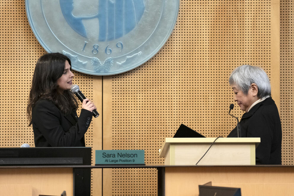 Two people at a dais, one with a microphone and the other with a book.