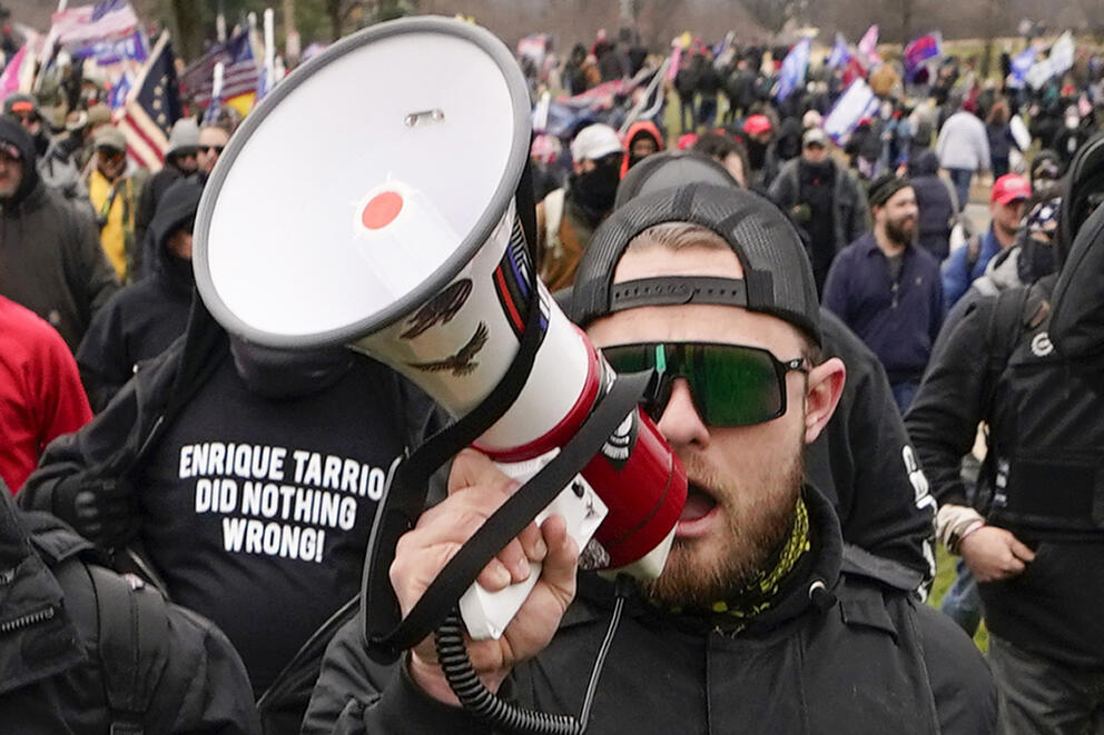 A person uses a megaphone in a crowd. A person in the background wears a shirt that says "Enrique Tarrio Did Nothing Wrong!" 