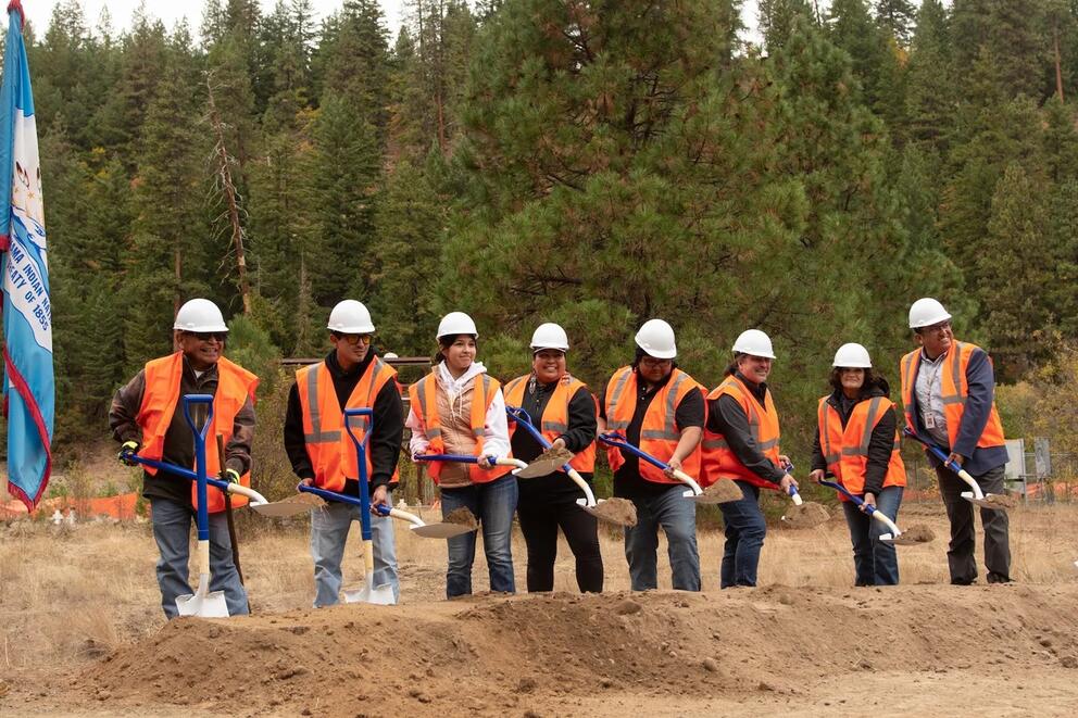 Eight people in hard hats and safety vests pose for a ceremonial groundbreaking