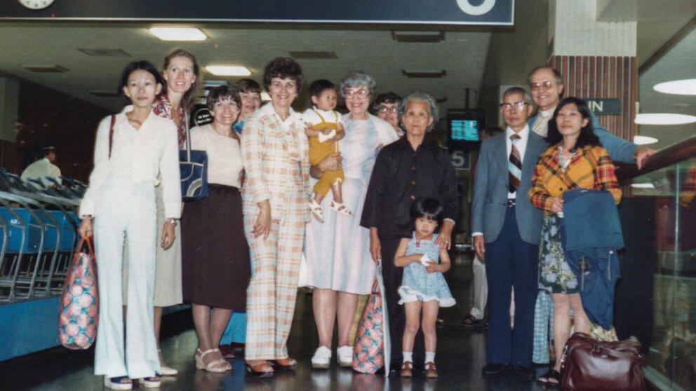 About a dozen people, aduults and children, stand in an airport lobby in 1980.
