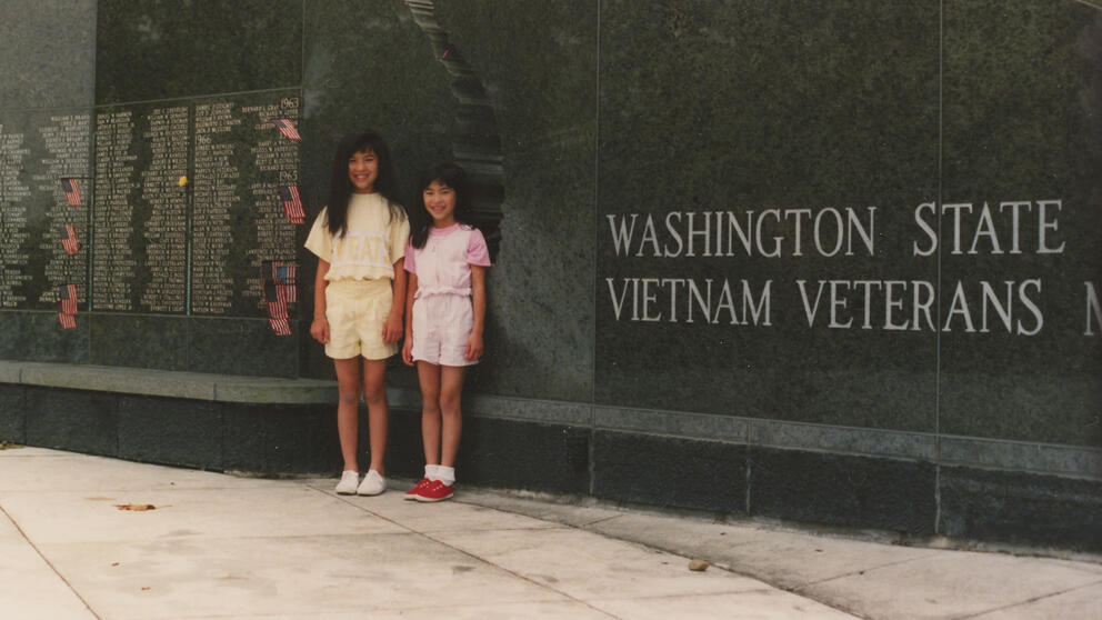 Two young girls stand in front of a large black marble wall with words inscribed on it.
