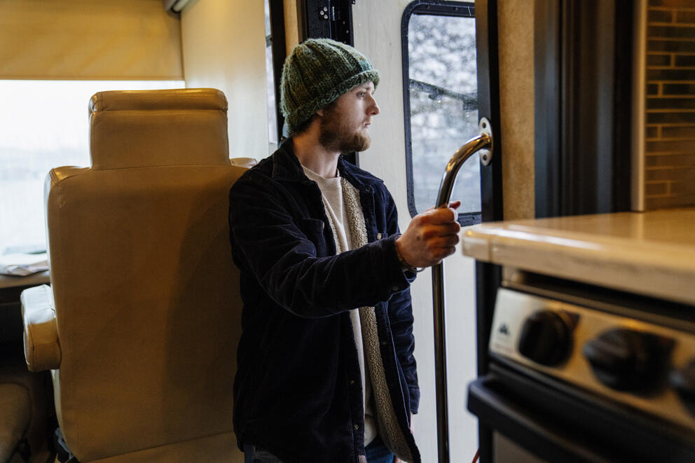 A man, wearing a beanie and fleece-lined coat, looks to the outside of an RV.