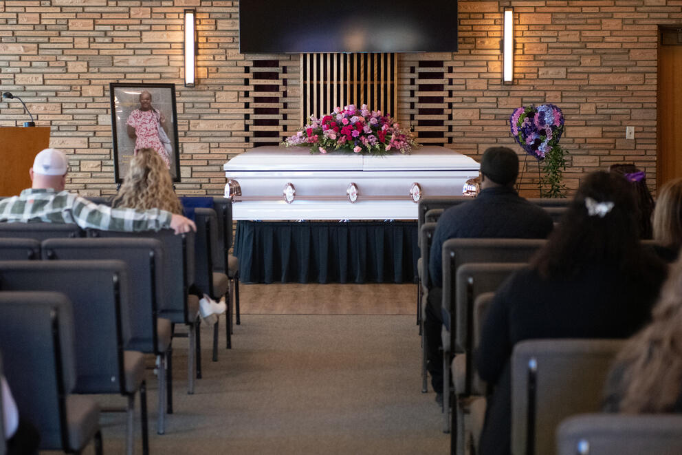 People in chairs look at a coffin at the front of a room decorated with flowers