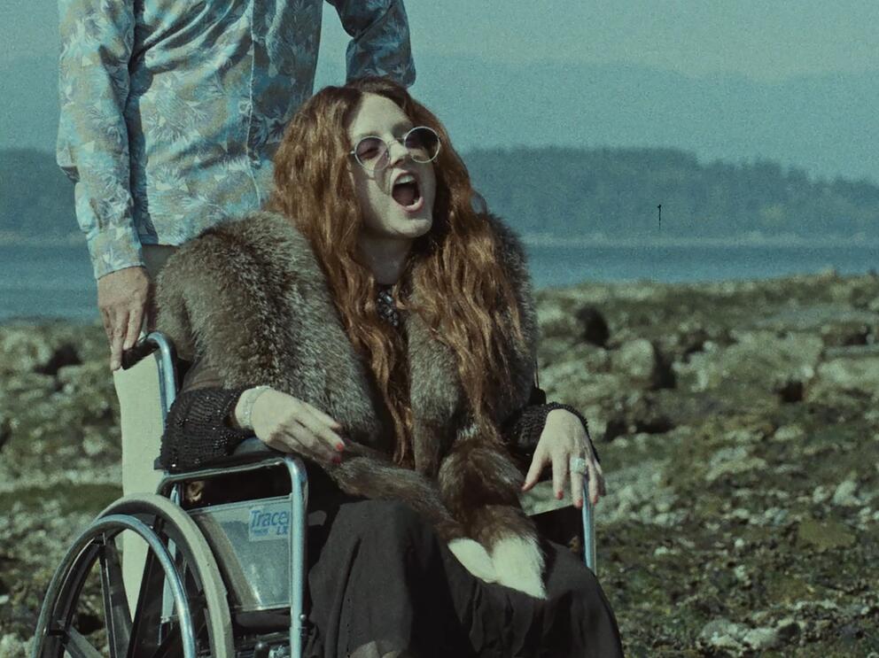 film still in which a woman with long red hair speaks from a wheelchair in an 1970s scene