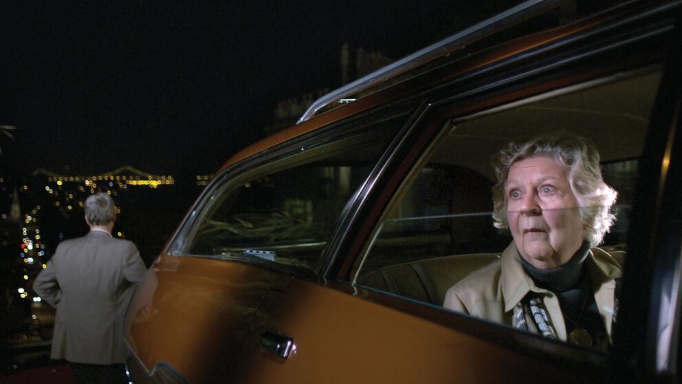 still from a movie in which an elder woman is looking out from a car window in alarm