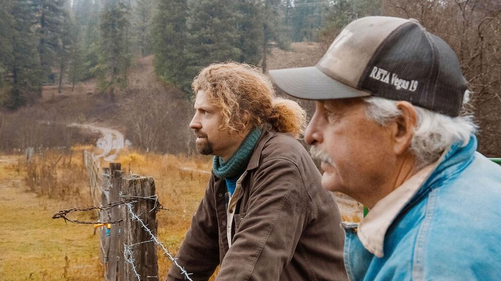 still image from a film with two men, one younger with long hair, one older in a cap, both looking at a field