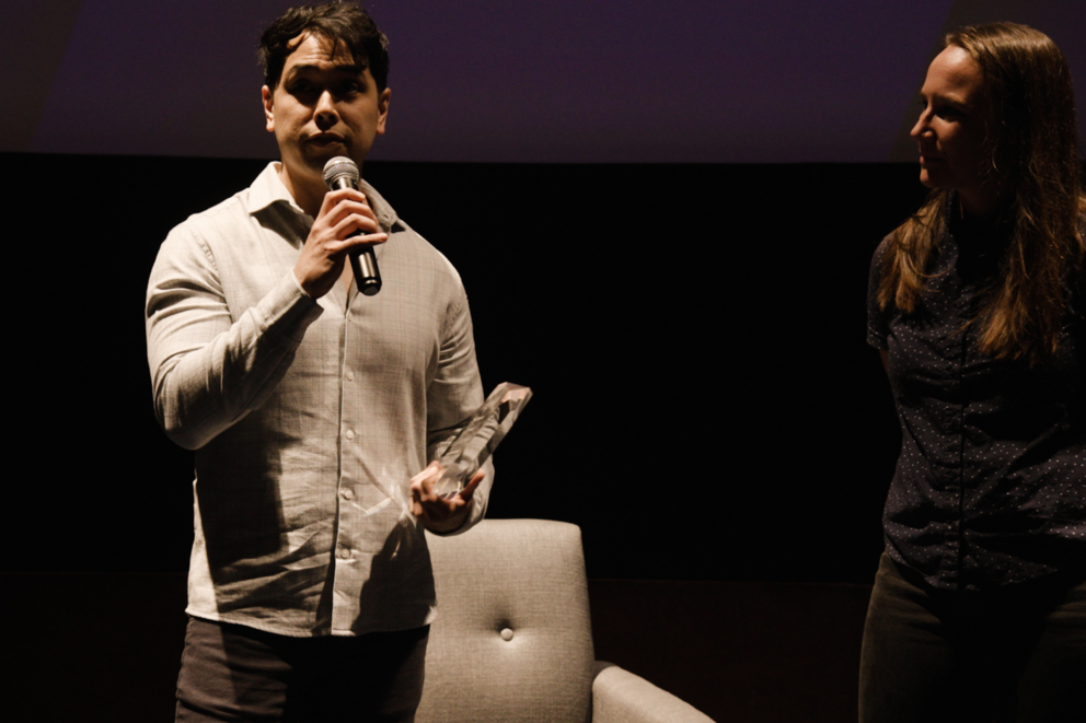 Andrew Inaba speaks into a microphone holding an award, Sarah Menzies stands to his right and looks his way