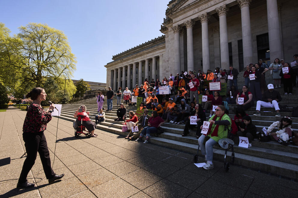 A person speaks on a microphone in front ofa  group of people who are on steps in front of a building with classical column. The people are holding signs.