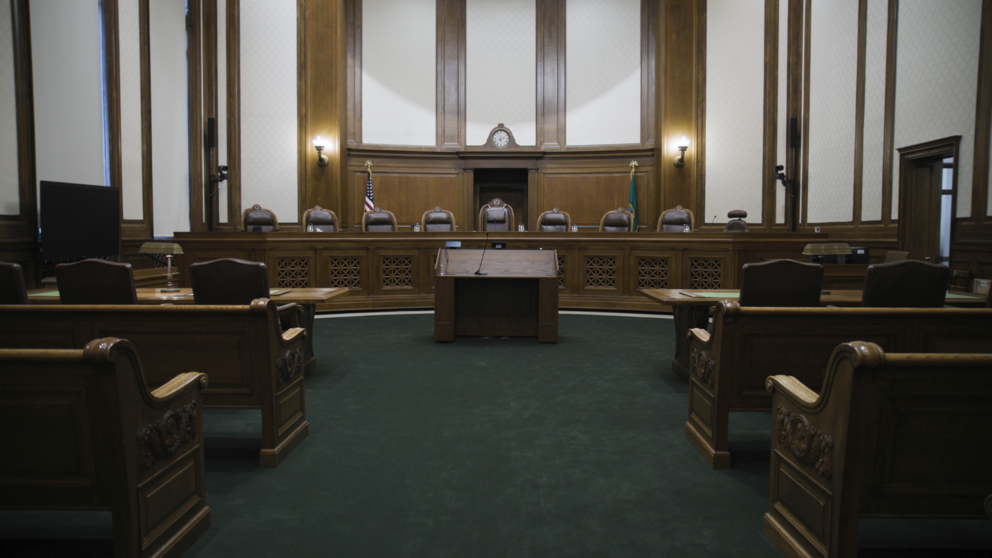 The washington state supreme court room with empty chairs behind the bench. All furniture is made of carved wood. The walls are white with wood trim, and the floor is green.