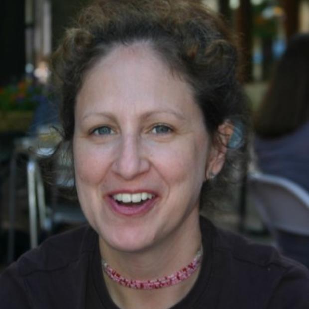 White woman with brown curly hair pulled into an updo wearing a beaded necklace and dark shirt smiles at the camera. She is sitting in a restaurant.