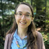 White woman with long, dark hair wearing glasses, a blue button-down shirt, a light brown cardigan and necklace with large blue and brown cylindrical beads smiles at the camera. There are trees in the background. 
