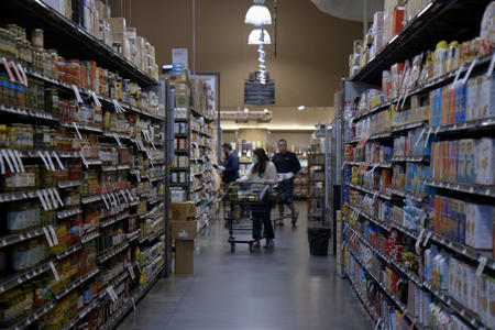 Three customers push carts through a mostly empty aisle in a grocery store.