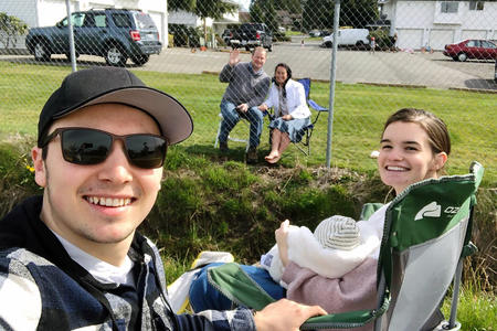 A young couple with a baby sit in camp chairs while grandparents sit on the other side of a chainlink fence