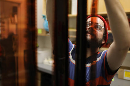 A man in glasses and a red hat reaches up out of frame to hang strips of 35mm film