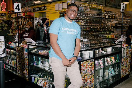 a man in an aqua colored shirt and light colored pants leans back against a glass display case full of marijuana products