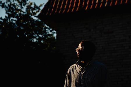 A man with glasses stands in shadow with a sliver of light hitting his left side