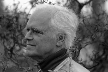 a man with white hair and wearing a dark turtleneck looks into the woods