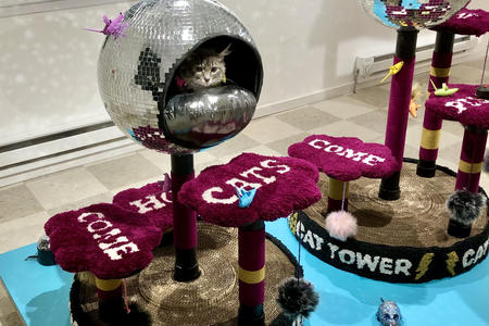 a gray cat peeks out from a cat tower sculpture shaped like a mirrored disco ball 