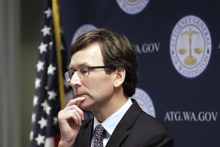 A man wearing glasses and a suit faces away from the camera, holding his chin in his hand. There is a flag behind him. 
