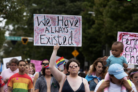 A person in a crowd holds a sign that reads "We have always existed."