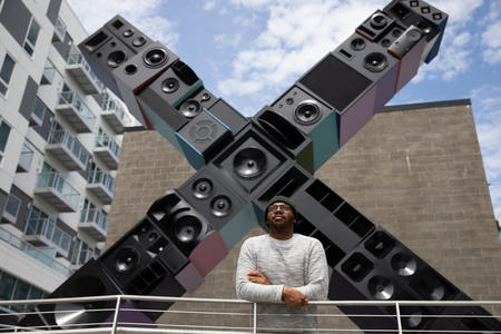 Person in hat in front of large 20 by 20 foot X sculpture made out of stacked boxes that look like speakers