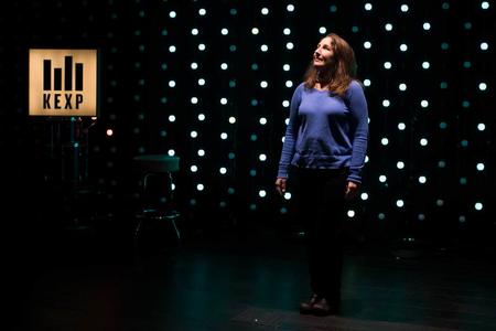 KEXP DJ Cheryl Waters standing in the dark KEXP studios with blue lights behind her. 