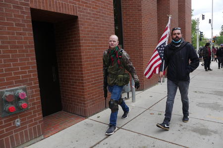 Two men walk down the street, one with an AR-15 and the other with a gun