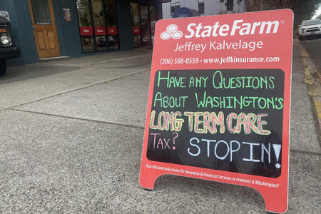 A red State Farm sandwich board with a chalk message, "Have any questions about Washington's Long Term Care Tax? STOP IN"