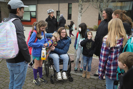 A group of parents and children group together on the sidewalk outside their school.