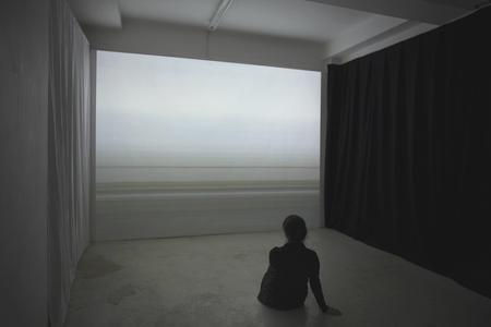A woman sitting on concrete floor looking out a wall-sized window into dull ocean/sky horizon