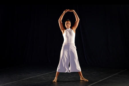 a woman in a white dress stands on a black stage with arms overhead