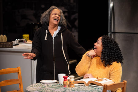 Two people on a stage, one person sits at a table, she is wearing a yellow shirt and has dark hair. The person she is looking at, who is standing, is dressed in dark clothing and has grey hair.