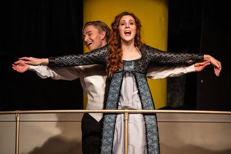 Woman in a white and blue dress stands with arms stretched out, a man standing behind her. They both appear to be singing.