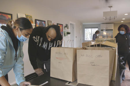 a group of masked people stand around a countertop with brown grocery bags on it