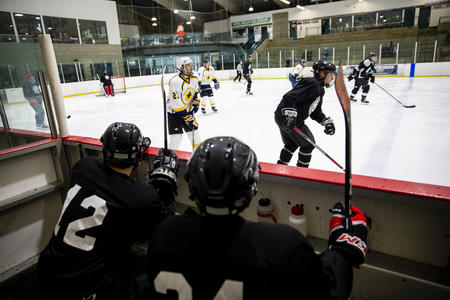 A hockey player on the sidelines looks on as two teams skate in the rink