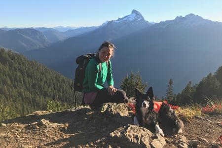 a person in a green jacket smiles on the top of a hill, with mountains in the background and a dog in the foreground