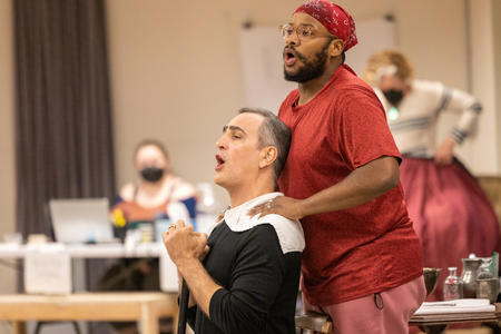 photo of a backstage rehearsal a man sits in front of another standing man, both singing