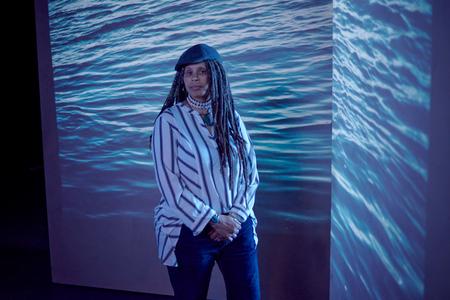 Portrait of Keith in a striped shirt, hat and dreadlocks with a photo of the ocean projected in the background