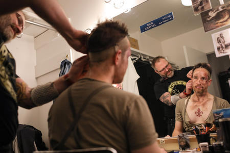 A wig is placed on a man with bloodied makeup and dirt on his face as he looks in the mirror
