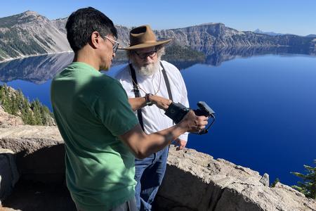 man in green shirt showing camera to man in overalls with a lake in background