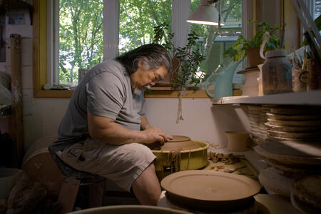 Reid Ozaki sits hunched over his spinning pottery wheel, as he molds clay into a platter